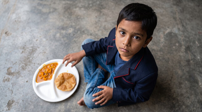 Poor young boy sitting on floor eating modest meal from styrofoam plate