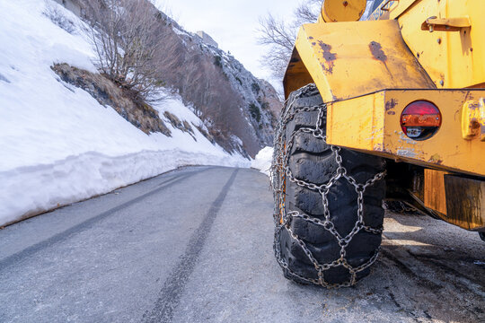 A yellow industrial vehicle wheel wrapped in metal snow chains stands on a paved mountain road beside melting snowbanks, a concept of winter road safety.