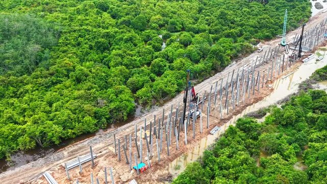 Aerial drone forward tracking shot over concrete foundation piles and operating piling rig machinery at a bridge and road construction site in a green landscape.