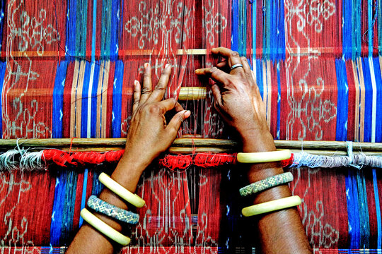 A top-down close-up view capturing the hands of an artisan weaving traditional Flores ikat textile featuring intricate patterns in vibrant shades of red, blue, and white. The weaver wears traditional 