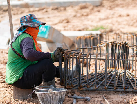 Female construction worker tying steel rebar cage with wire at a outdoor ground foundation site.