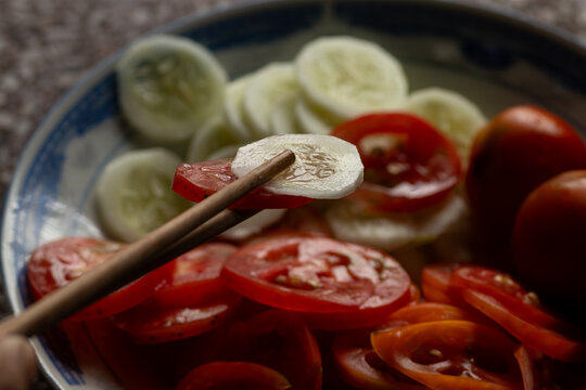 Wooden chopsticks picking up fresh sliced cucumber and juicy red tomato from a plate for a healthy vegan diet