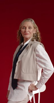 Vertical video: Mature woman posing in studio, turning to camera, leaning on stool in vest