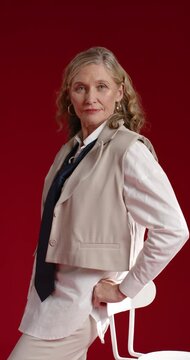 Vertical video: Senior woman adjusting pose on studio stool at photog cue for portrait, wearing tie