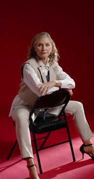 Vertical video: Woman in vest modeling after cue, adjusting pose on backward chair in red studio