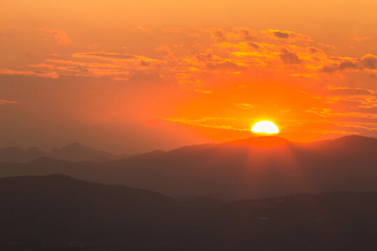 A vivid sunset with orange sky, sun half-hidden behind dark mountain silhouettes, creating a serene natural landscape.