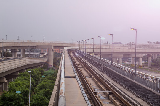 Elevated railway tracks run through an urban area, flanked by overpasses and streetlights, with greenery below under an overcast sky.
