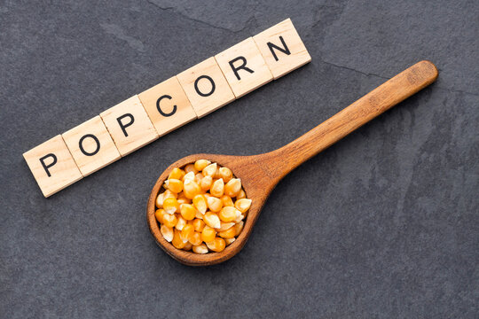 Popcorn kernels scattered on a dark surface with a wooden spoon and the word "POPCORN" spelled out with wooden blocks.