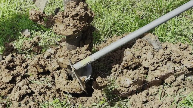 Close up of manual earth auger drilling into soil for post hole construction and foundation work in outdoor garden area.