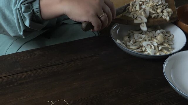 Hands chop fresh champignons with a knife on a cutting board on a wooden table