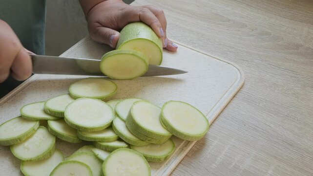 A woman's hand slices green zucchini into rings on a board