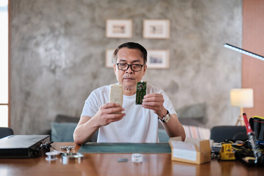 Mature Asian man checks and compares spare parts parcel, air conditioner remote control circuit board replacement, home electrical appliance fix, electronics component hobbyist, enthusiast workshop.