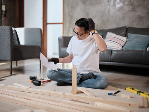 Asian mature man self-assembling flat-pack furniture with DIY tools and instruction guide, confused with wooden parts, home improvement, craft installation, hobby workshop, handyman active lifestyle.