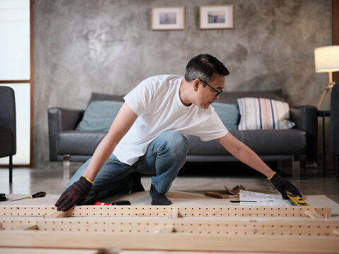 Asian mature man self-assembling wooden flat-pack furniture with DIY carpentry tools, following instruction manual, home improvement, craft installation, hobby workshop, handyman active lifestyle.