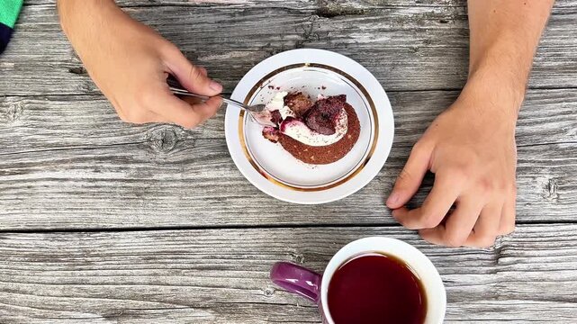 Top-down shot of a person eating a chocolate pastry with a fork next to a mug of hot tea on a rustic wooden table. Ideal for cafe menus, bakery ads, and food lifestyle media.