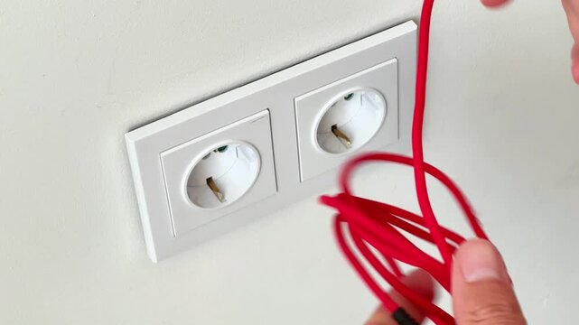 Close-up shot of hands holding a tangled red cable in front of double European wall power sockets. Ideal for tech troubleshooting, home electronics ads, and cable management media.