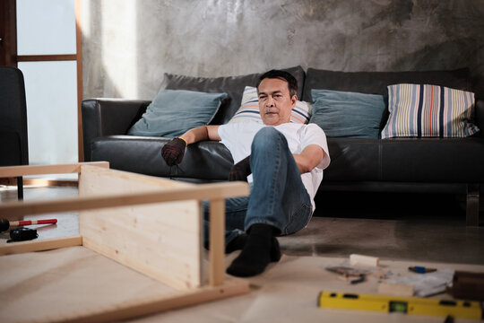 Asian mature man self-assembling carpentry furniture with DIY tools, disheartened from difficulty, tired and resting at living room sofa, home improvement, craft installation, hobby wooden workshop.