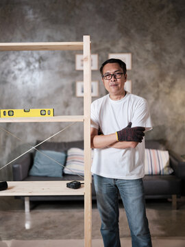Asian mature man standing near self-assembling wooden furniture with DIY tools, strength achievement satisfied, cheerful and proud home improvement accomplishment, hobby carpentry workshop handyman.