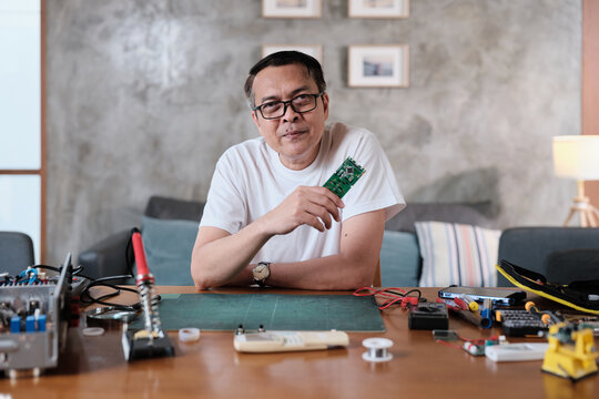 Mature Asian man showing pride in circuit board after successful soldering job, multimeter check, cheerful job achievement, home electronics appliance repair handyman, DIY fix tools, hobby workshop.