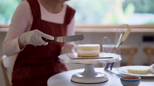 Asian baker spreading cream frosting on layered cake with spatula in bright home bakery kitchen