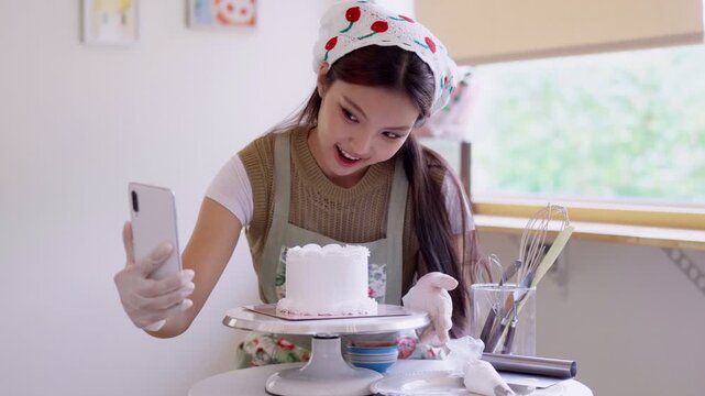 Young Asian baker filming smartphone video of finished white cake on stand in bright home kitchen