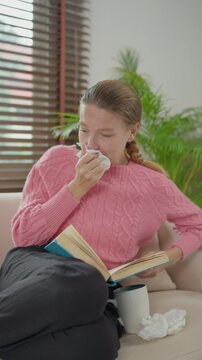 Sick woman sneezing while reading book on couch