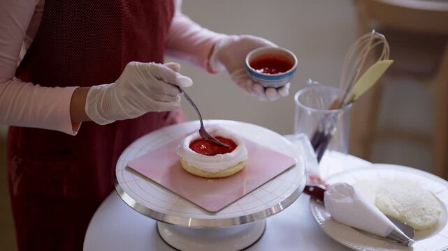 Professional Asian baker spreading red fruit topping on white frosted cake while preparing dessert on stand