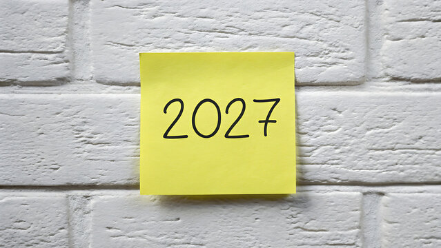 Yellow sticky note with 2027 written on it pinned to a grey wall