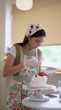 Young Asian baker placing red cherries on white frosted cake in bright home bakery kitchen