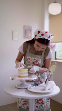Young Asian baker assembling layered cake with pastry spatula on rotating stand in bright home studio kitchen