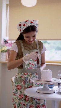 Young Asian baker smoothing icing on white layered cake with spatula in bright home kitchen