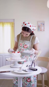 Young Asian baker decorating white layer cake with fresh cherries in bright home kitchen studio