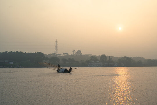 A small fishing boat with two fishermen casting a net on a calm river during a golden sunset in a rural landscape
