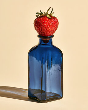 Fresh red strawberry balanced on a vintage blue glass bottle against a warm cream background, creating a colorful surreal food still life with bold contrast.