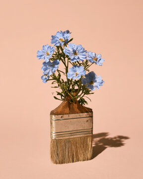 Blue flowers growing from a vintage paintbrush against a soft peach background, creating a whimsical surreal concept combining painting, nature and artistic creativity.