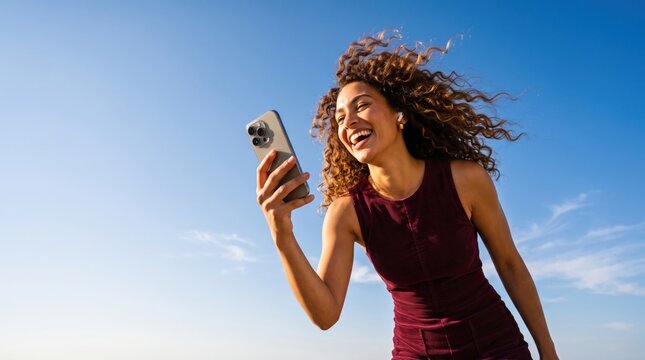 Happy Young Woman with Curly Hair Using Smartphone Outdoors Under Blue Sky.