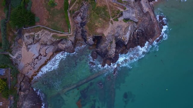 Top-down drone shot moving away from a rocky shoreline, revealing turquoise water, breaking waves, coastal paths and a small seaside house.