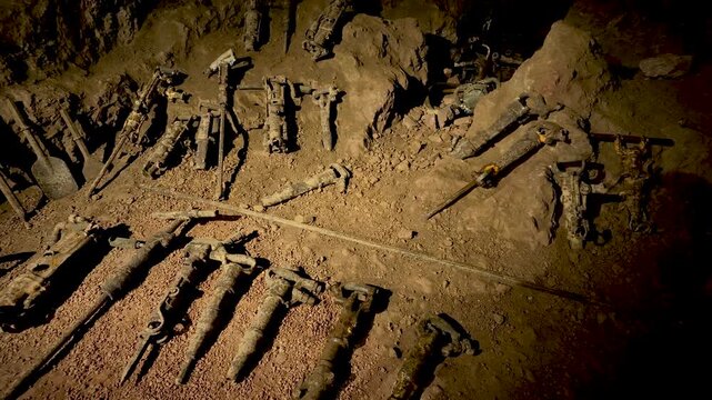 Rows of old pneumatic rock drills lie on the dusty mine floor as the shot slowly pans across the abandoned equipment.
