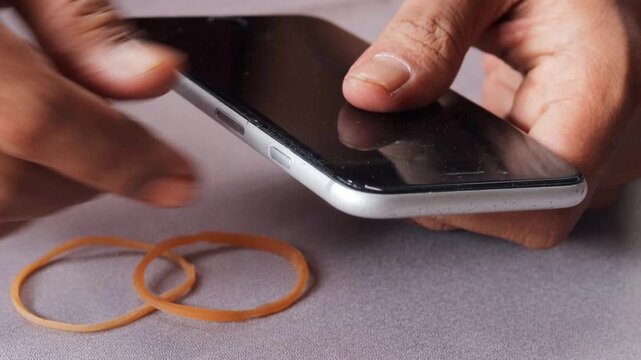 Close-up of Asian man's hands securing a slightly lifted smartphone screen with a rubber band as a temporary repair. Phone maintenance, financial hardship, and extending device use.