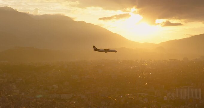 Kathmandu, Nepal, drone racking shot of a passenger plane on approach to Tribhuvan International Airport, passing directly across a bright golden hour sun setting behind the valley mountain