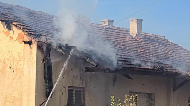 Firefighters extinguish burning house roof with water hose.