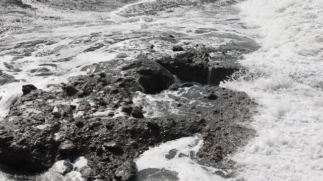 Waves crash against rugged, wet rocks in monochrome. Foam swirls, revealing hidden crevices beneath the spray. Surf churns, eroding the coastline with persistent force