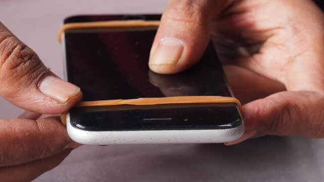 Close-up of Asian man's hands securing a slightly lifted smartphone screen with a rubber band as a temporary repair. Phone maintenance, financial hardship, and extending device use.