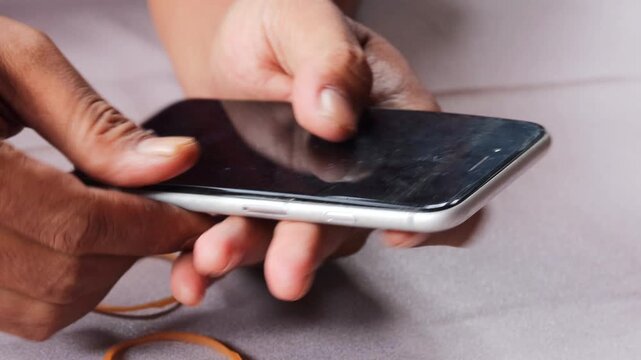Close-up of Asian man's hands securing a slightly lifted smartphone screen with a rubber band as a temporary repair. Phone maintenance, financial hardship, and extending device use.