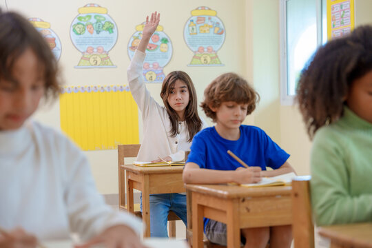 Diverse elementary students studying in classroom girl raising hand to answer teacher question