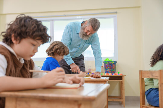 Senior school teacher explaining tasks and pointing at notebooks of elementary students studying inside a bright classroom