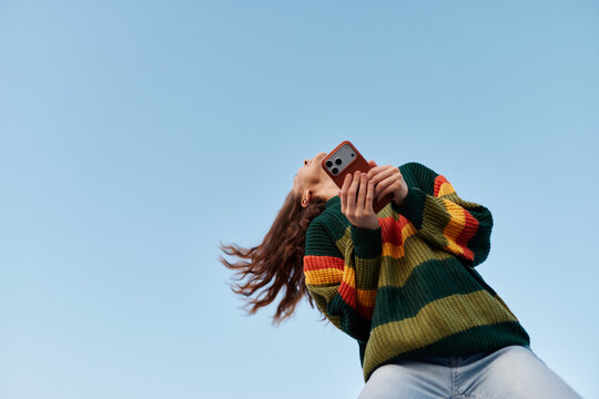 Low angle view of a young woman in a colorful striped sweater photographing with her smartphone against a clear blue sky. Concept of mobile photography and freedom