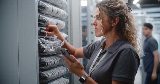 Female IT Specialist Checks Ethernet Connections and Verifies Rack Topology on Smartphone, Matching Physical Ports With Digital Records While Male Technician Works on Another Server Cabinet.