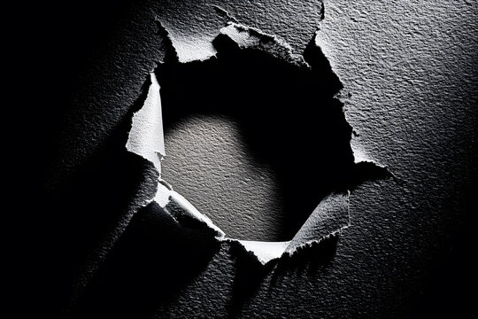 Hole breaking through black damaged wall paper on black background. Hole in black paper or with torn edges and piercing sunlight and rays through it. Broken paper wall art.