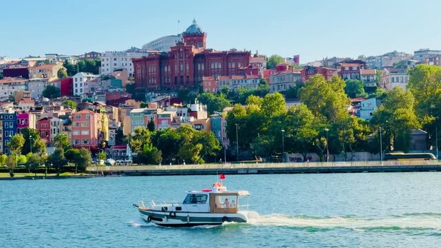 Small motor boat cruises on Golden Horn bay water against colorful Balat district houses in Istanbul, Turkey. Scenic urban view with ancient Phanar red brick college under a blue summer sky.
